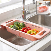 Pink plastic adjustable over-sink drain basket with broccoli, tomatoes, and bell peppers in a kitchen sink.
