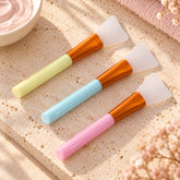 Three silicone face mask brushes with pastel handles in yellow, blue, and pink, on a textured stone surface with a bowl of