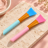 Two silicone face mask brushes, one pink and one blue, rest on a light marble surface.
