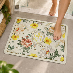 A person's bare feet step onto a rectangular bath mat featuring a botanical design with yellow, pink, and white flowers on a