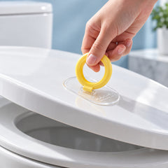 A hand lifts a white toilet seat with a yellow ring and clear base, highlighting the simple design.
