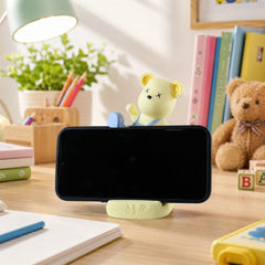 A light yellow bear phone stand holding a black smartphone horizontally on a bright wooden desk.