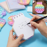 A person's hands writing on a pastel hexagon pop it fidget notebook with a black pen on a blue surface.