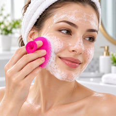 A woman with a white towel on her head is washing her face with a pink silicone cleansing brush, creating white foam.