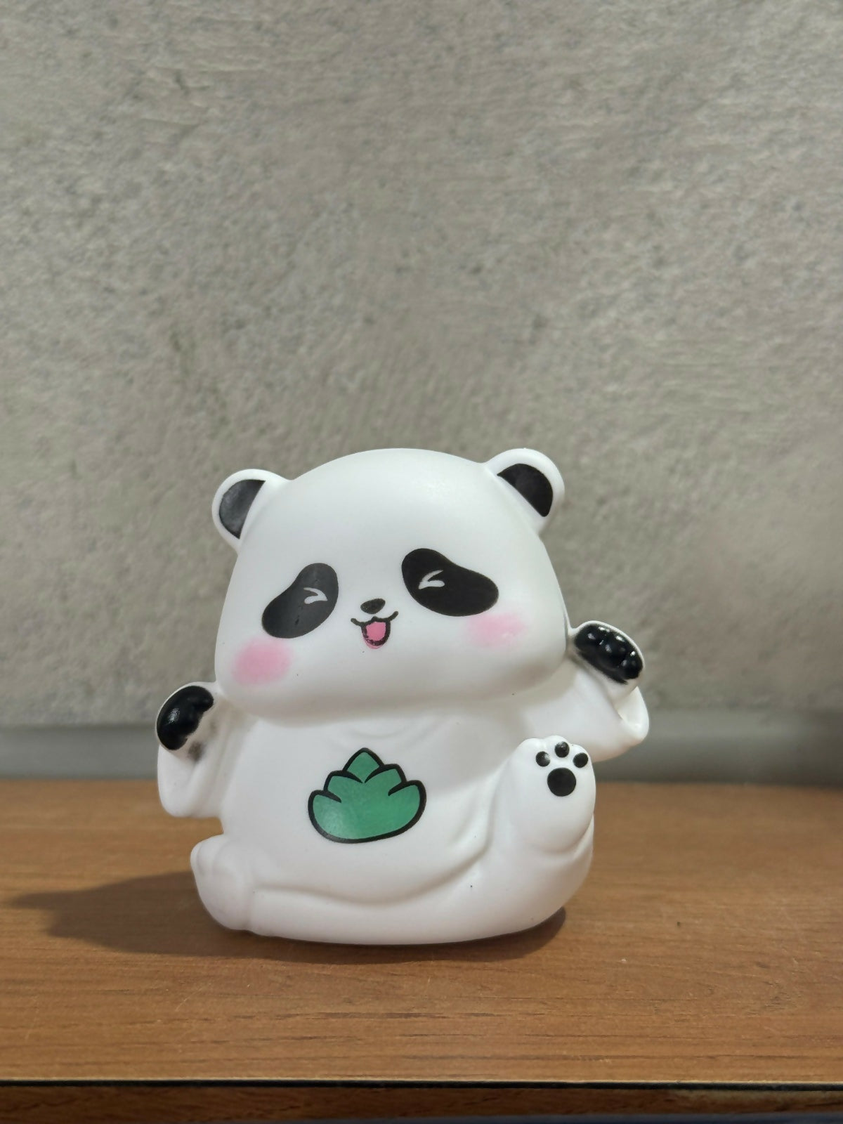 A white panda night light with black ears, eye patches, and a green leaf design on its stomach, resting on a wooden surface