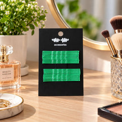 A black card with green metal bobby pins stands on a wooden vanity, surrounded by a plant, perfume, and a small dish with