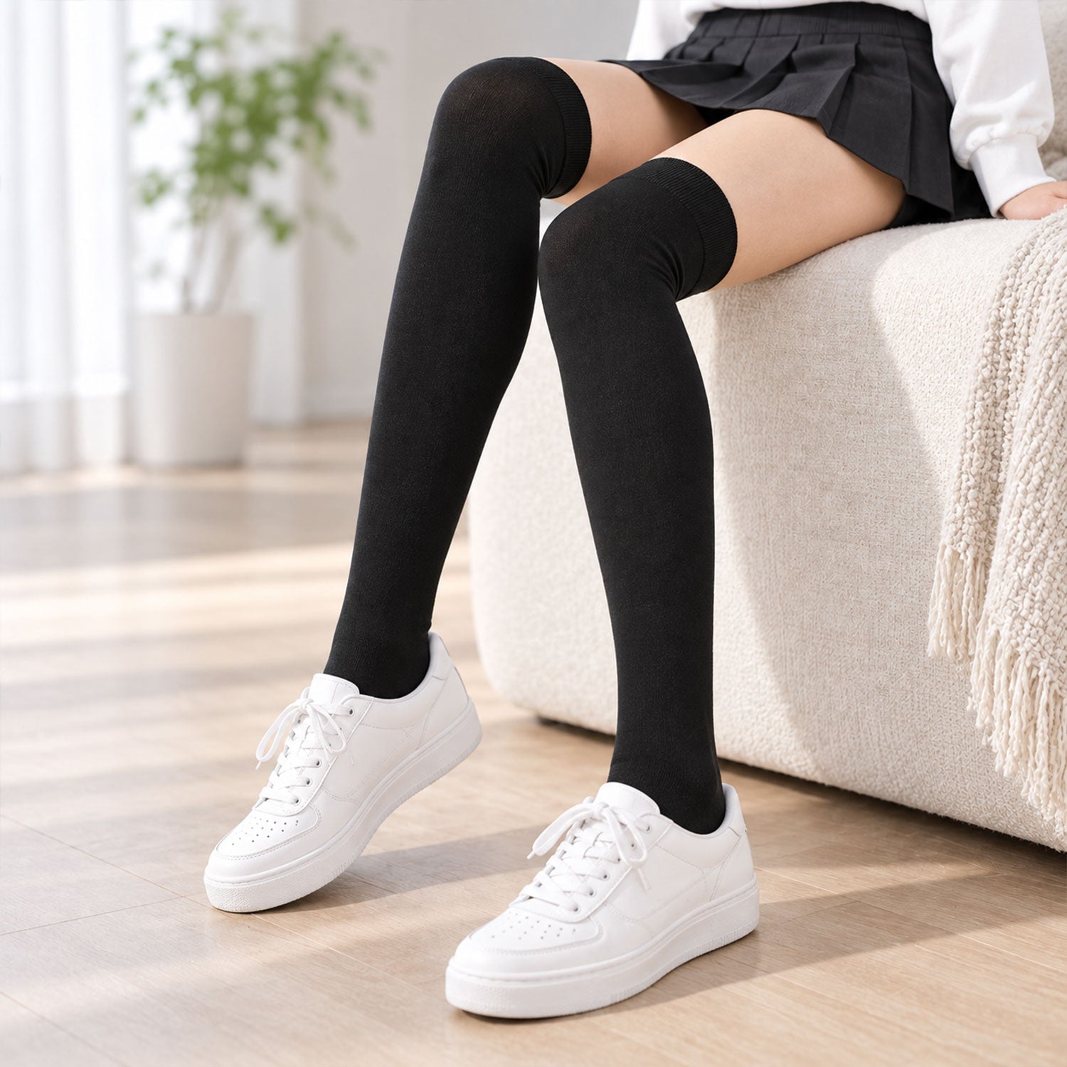 A person sitting on a sofa, wearing black over-knee socks and white sneakers on a light wooden floor.