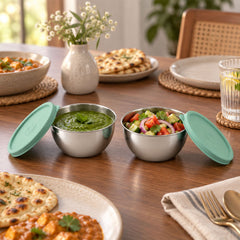 Two open stainless steel bowls with green lids, filled with green chutney and salad, on a wooden dining table.