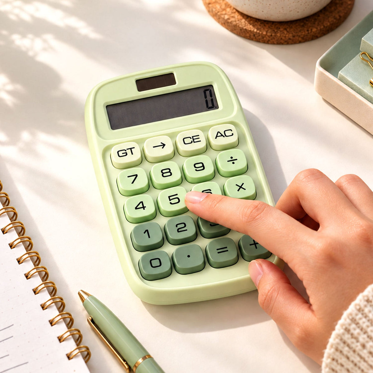 A person's hand presses a key on a light green calculator with dark green number buttons, next to a notebook and pen.
