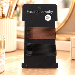 A black card holds two rows of brown and black metal bobby pins, displayed on a vanity with a mirror.