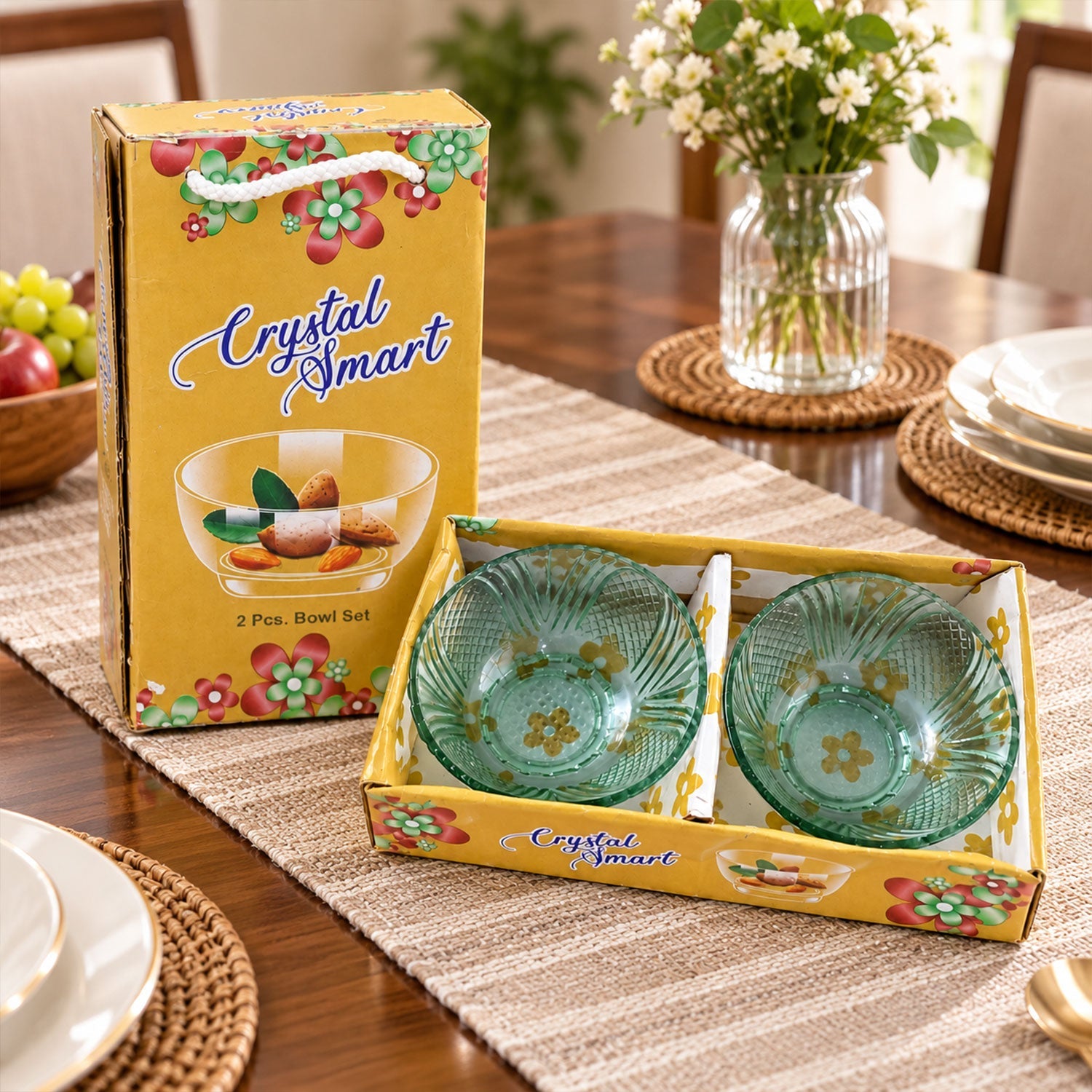 Two green glass bowls in a yellow box, with a vase of flowers and fruit on a wooden table.
