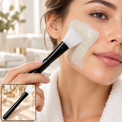 A woman applies a light green face mask to her cheek with a black and white silicone brush. Her hair is tied back, â€“ view 3