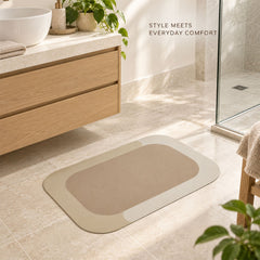 A rectangular bath mat with rounded corners, featuring a beige center and a light cream border, rests on tiled floor in a