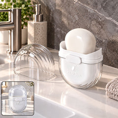 A clear and white oval soap box with a bar of soap inside, displayed open on a bathroom counter. The clear lid is â€“ view 3