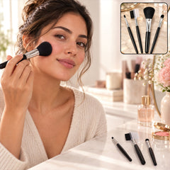 A woman applies blush with a large black and silver makeup brush, with three other brushes and a mirror on the table.