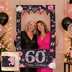 Two smiling women holding a black and pink 60th birthday photo booth frame, surrounded by pink and black balloons.