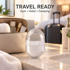 A closed clear and white oval soap box on a bathroom counter, with a travel bag and suitcase in the blurred background.