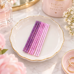 Nine glitter bobby pins in shades of pink and purple are arranged on a white scalloped dish with gold trim.