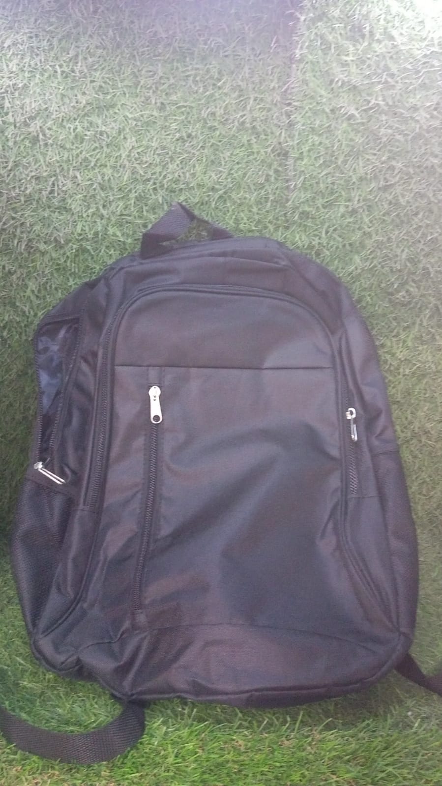 A black fabric backpack with multiple zippered compartments and side pockets lies on a green grassy surface.