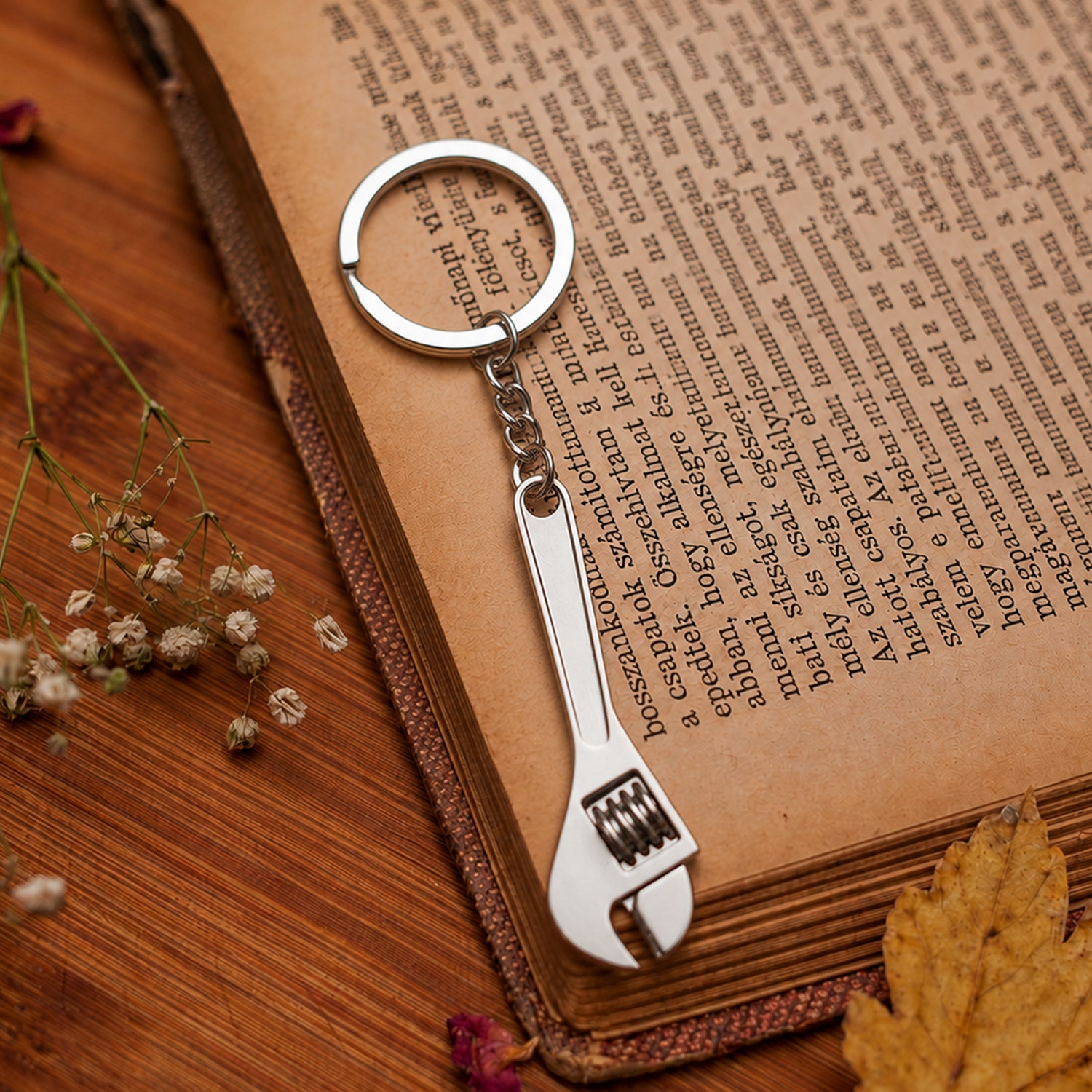 A silver metal mini adjustable wrench keychain lies on an open book with aged pages and small dried flowers.