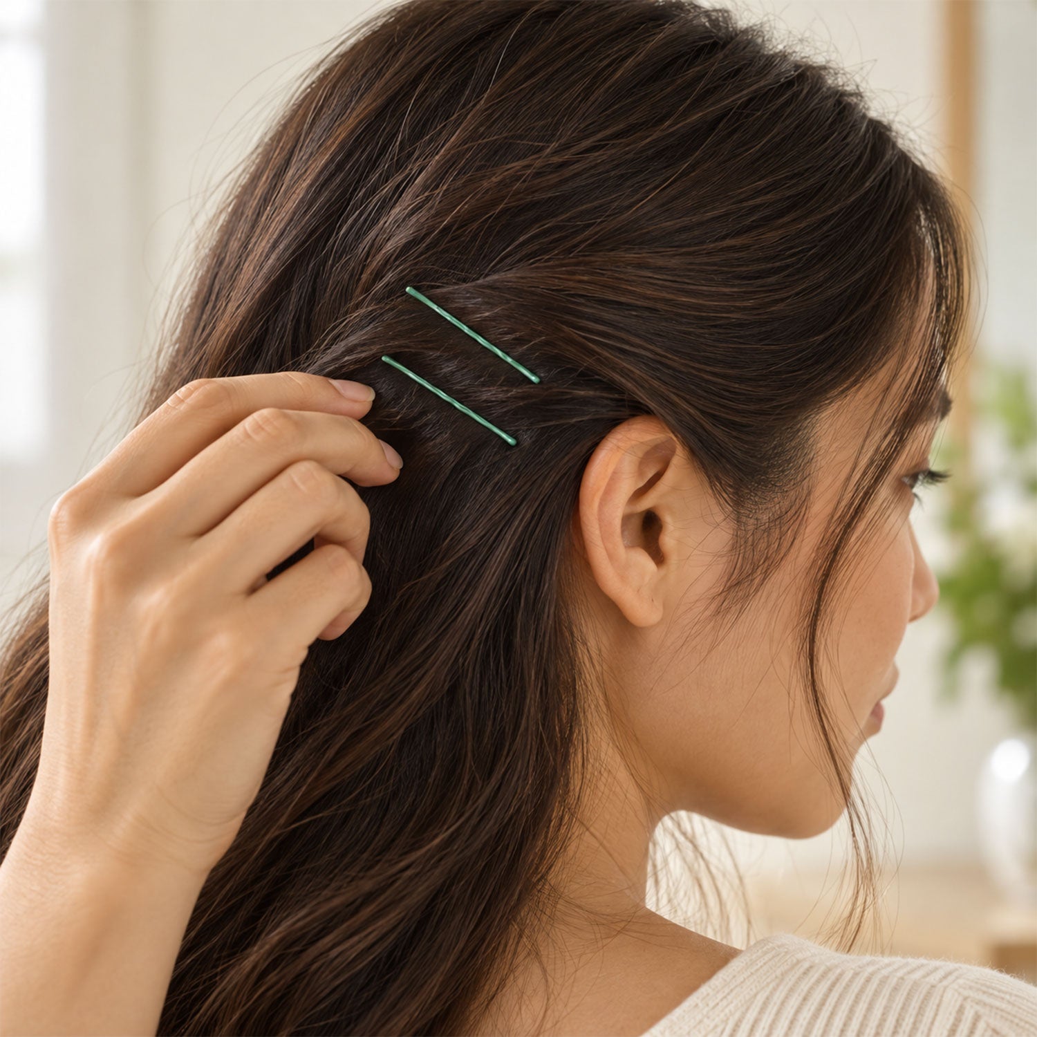 A close-up view of a woman's dark hair with two green bobby pins visible, as she gently touches her hair near her ear.