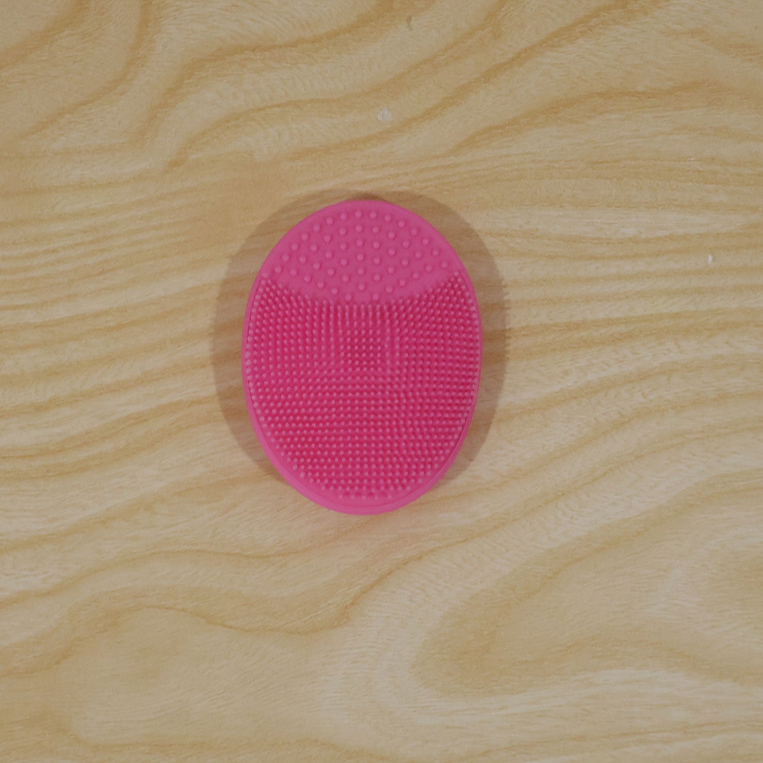 A single pink oval silicone cleansing brush with varied bristle textures rests flat on a light wood grain surface.