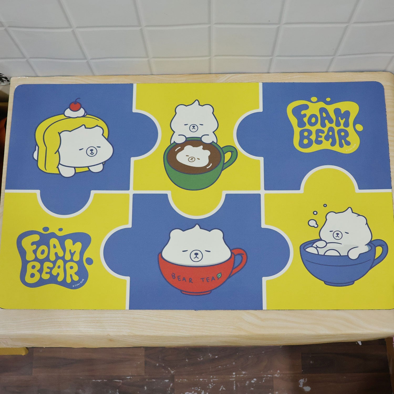 A blue and yellow bath mat with cartoon bears in cups is placed on a light wooden surface.