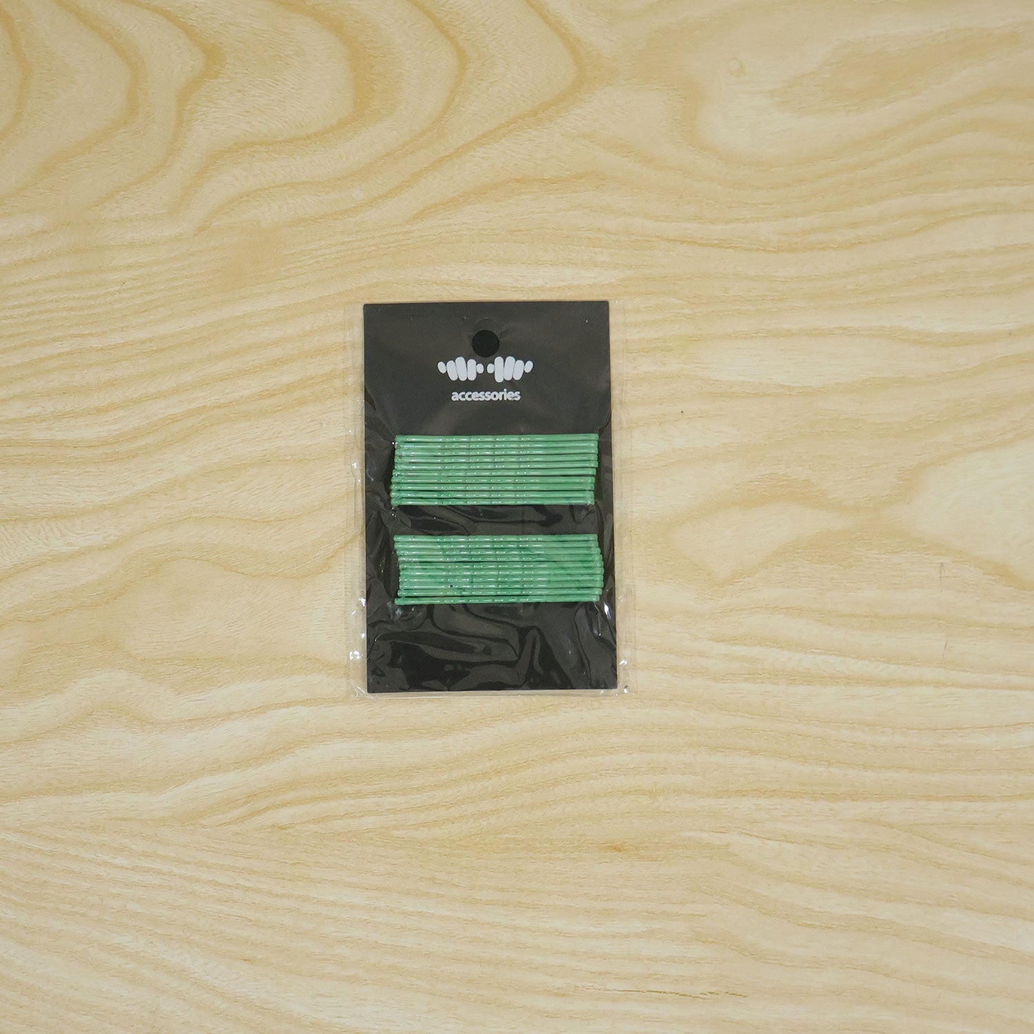 A black card with green metal bobby pins is sealed in clear plastic packaging, placed centrally on a light wooden surface.