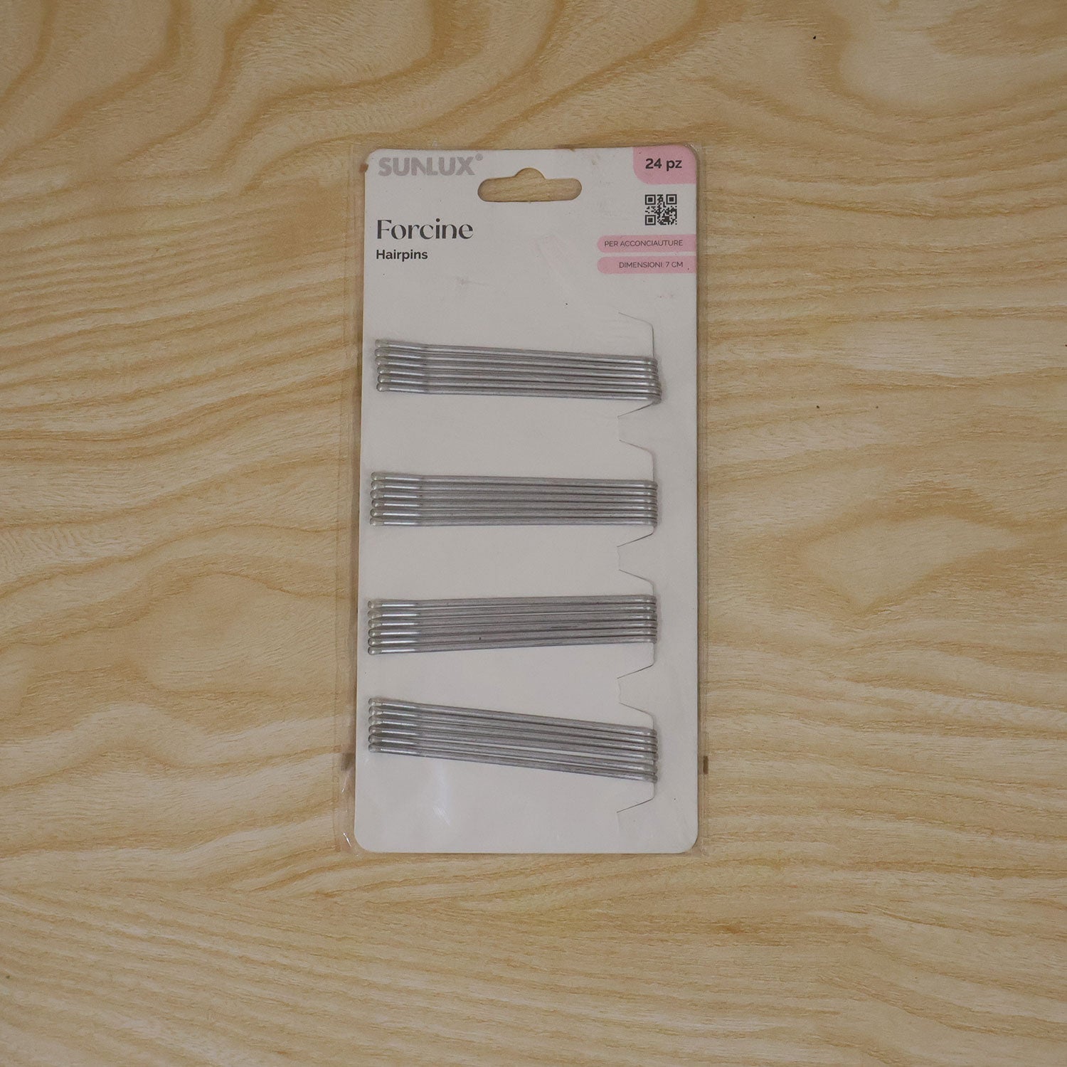A white package of twenty-four silver hairpins displayed flat on a light brown wooden surface.