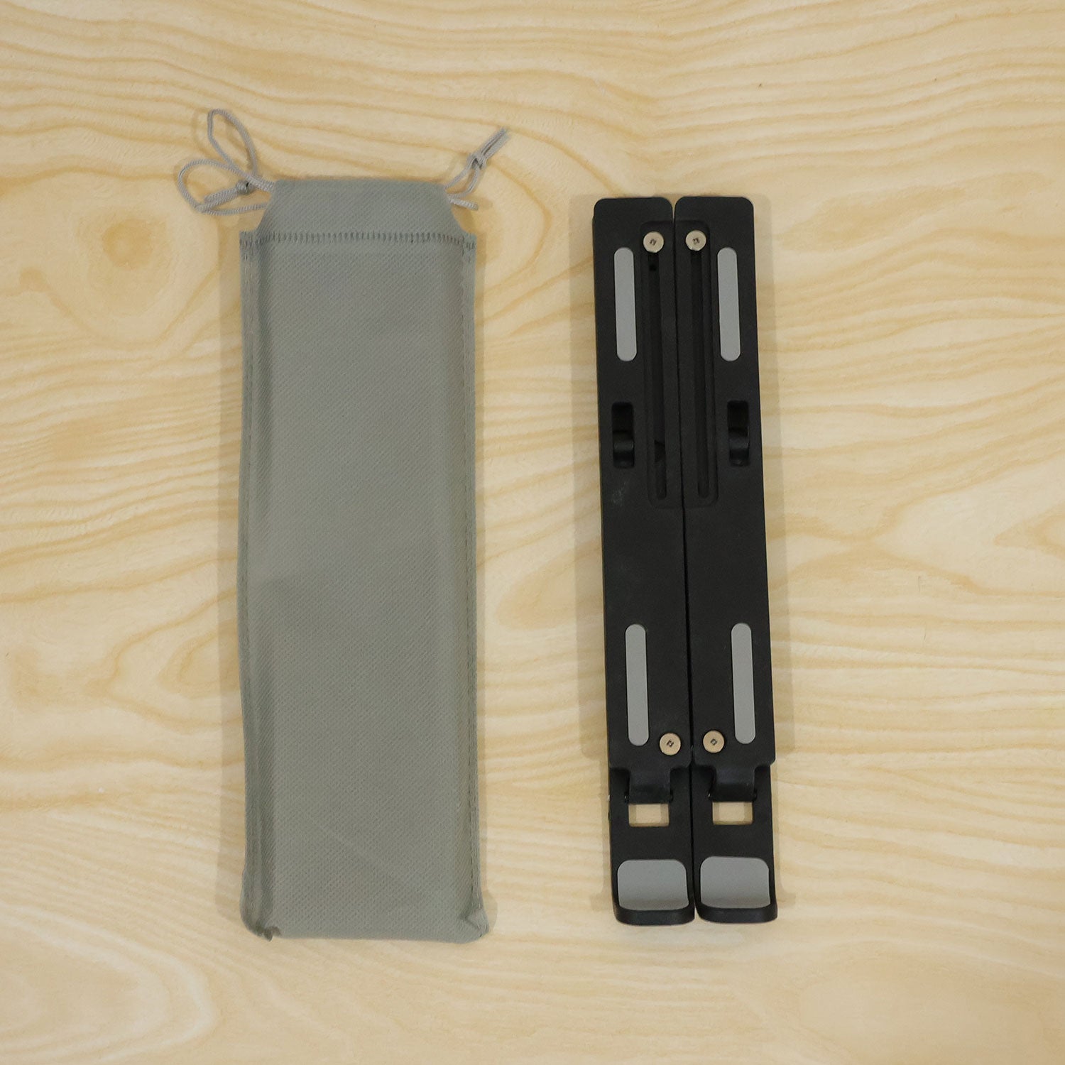 A folded black laptop stand with grey pads lies beside a light grey drawstring carrying bag on a wooden surface.