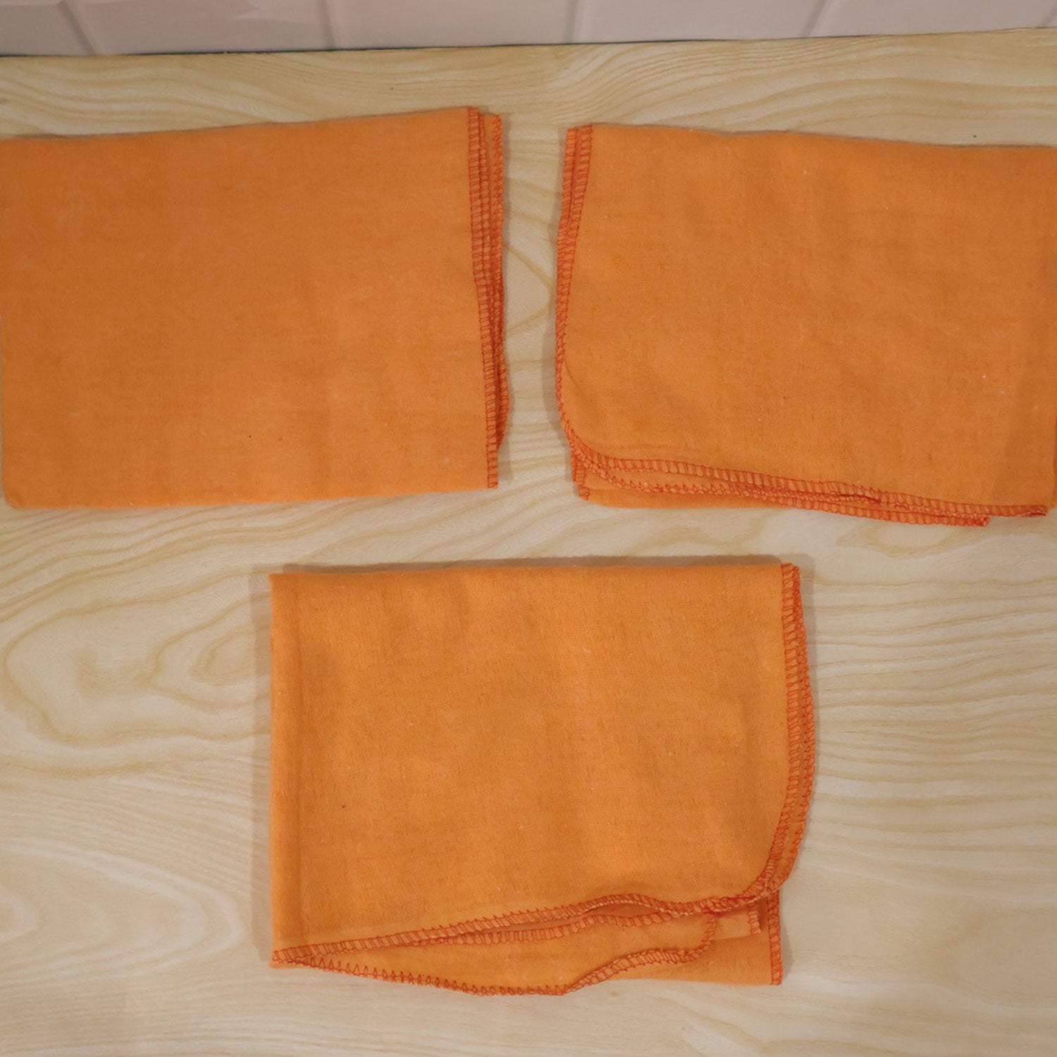 Three orange cleaning cloths are arranged on a light wooden surface, one unfolded and two folded.
