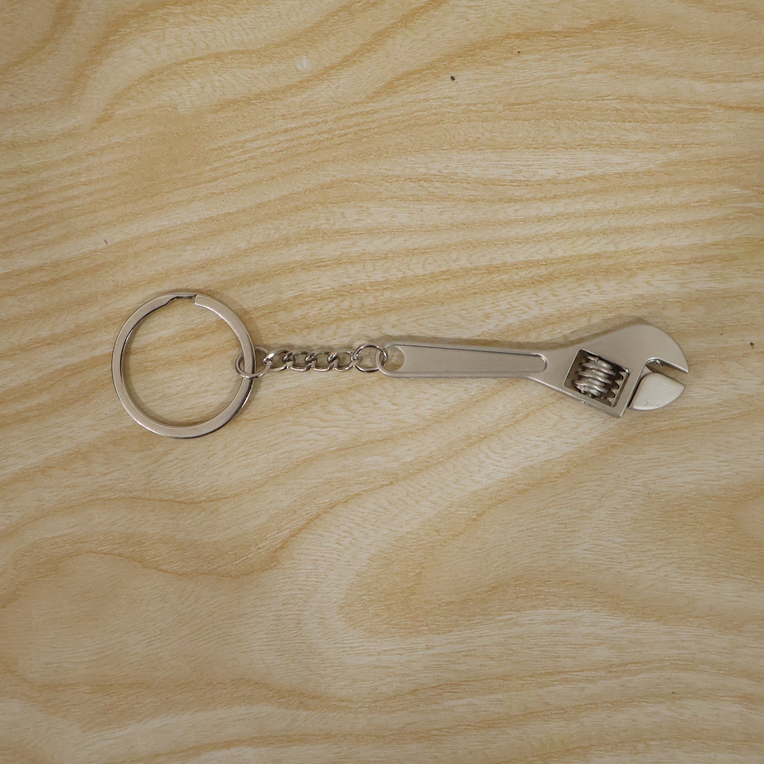A silver metal mini adjustable wrench keychain with a key ring and chain is laid flat on a light-colored wooden surface.