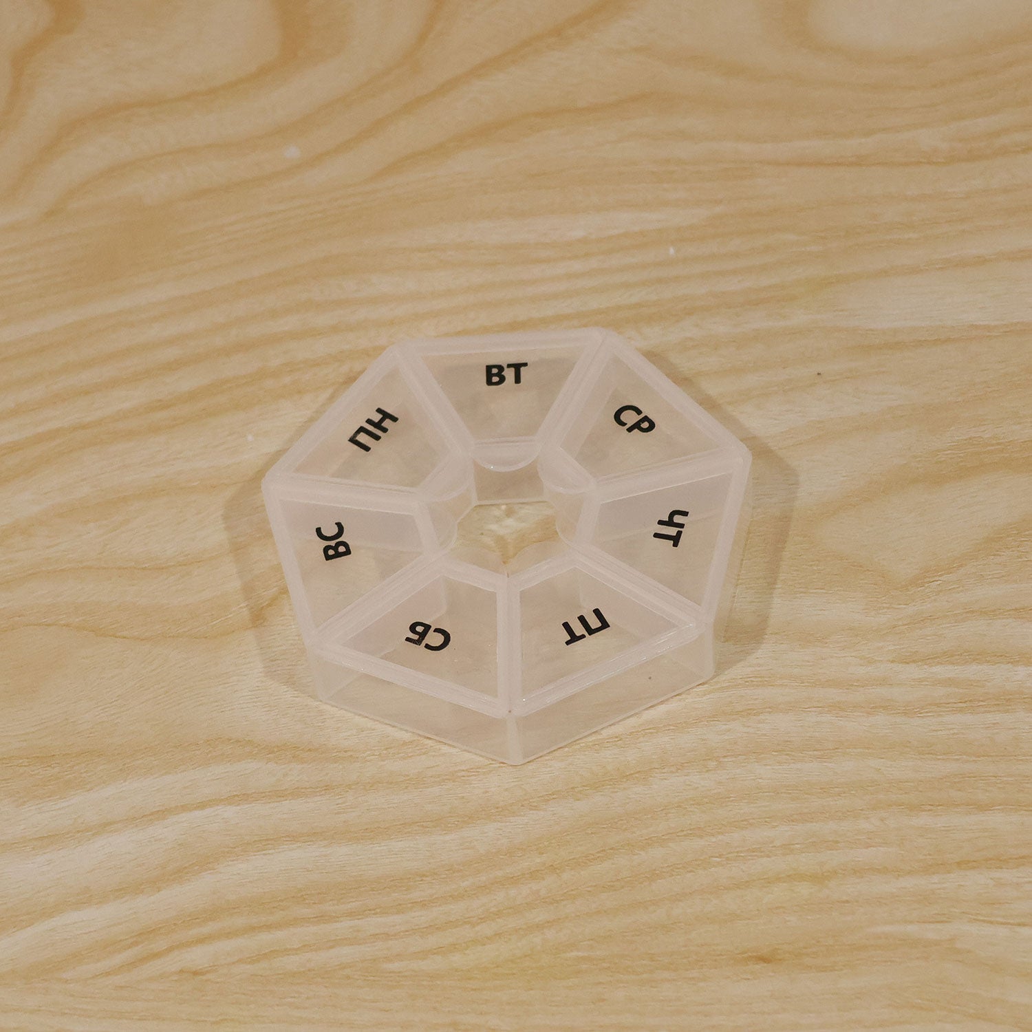 A clear hexagonal pill organizer with seven labeled compartments rests on a light wooden surface.