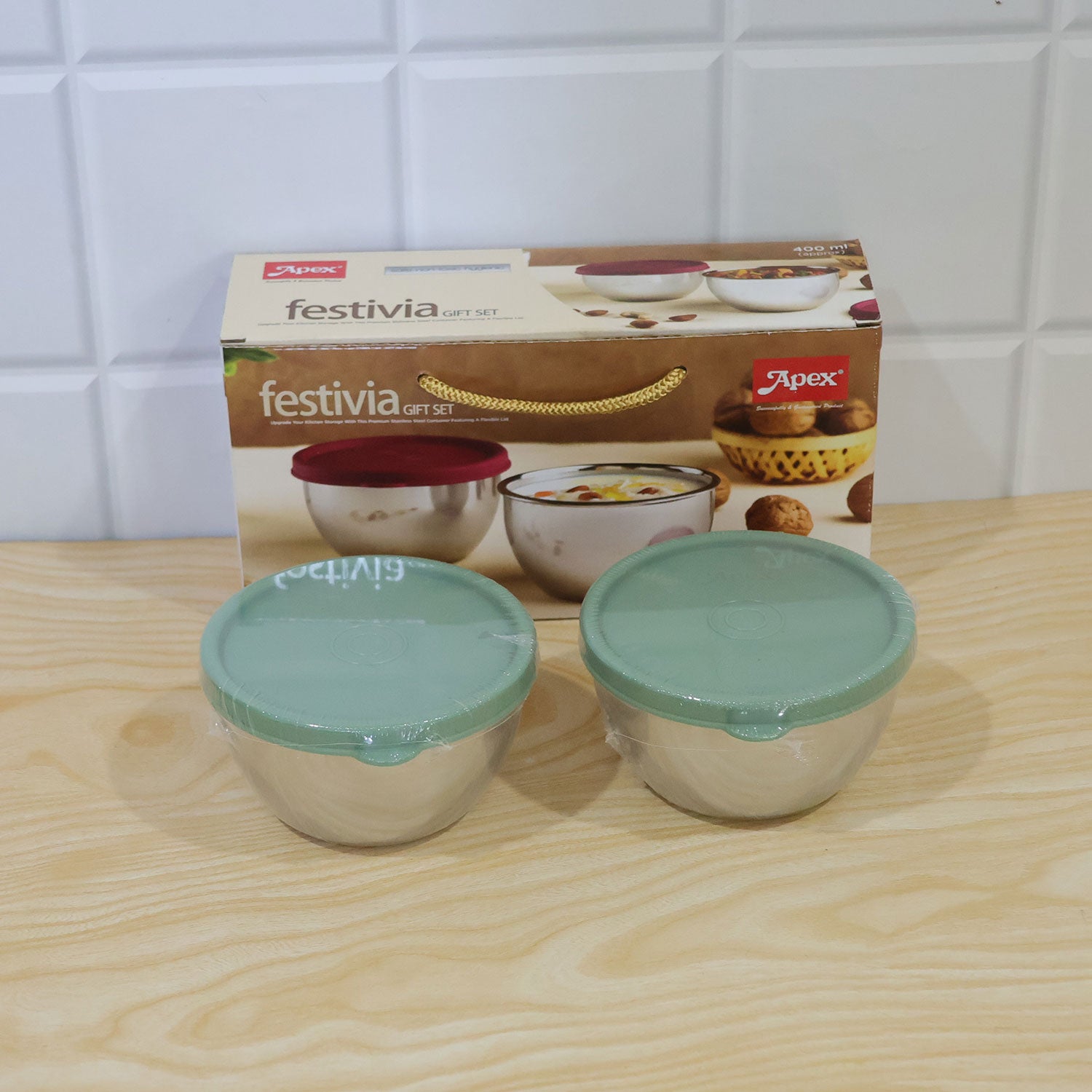 Two new stainless steel bowls with green lids, still in plastic wrap, in front of their product box on a wooden surface.