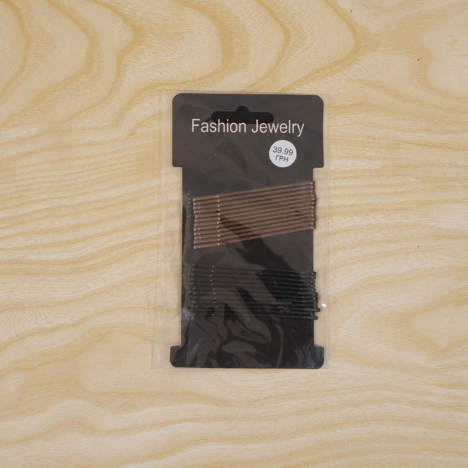 A black card with brown and black bobby pins is sealed in clear plastic, resting on a light wooden surface.