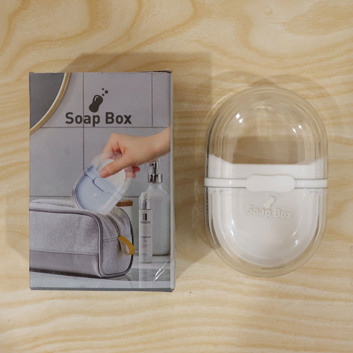 A clear and white oval soap box next to its gray and white product packaging box on a light wooden surface.