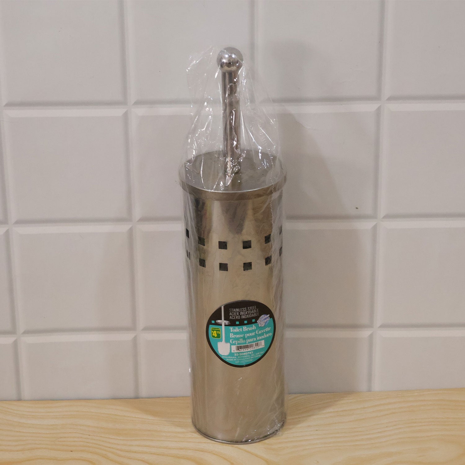 A stainless steel toilet brush and holder set, wrapped in clear plastic packaging, standing on a light wooden surface.