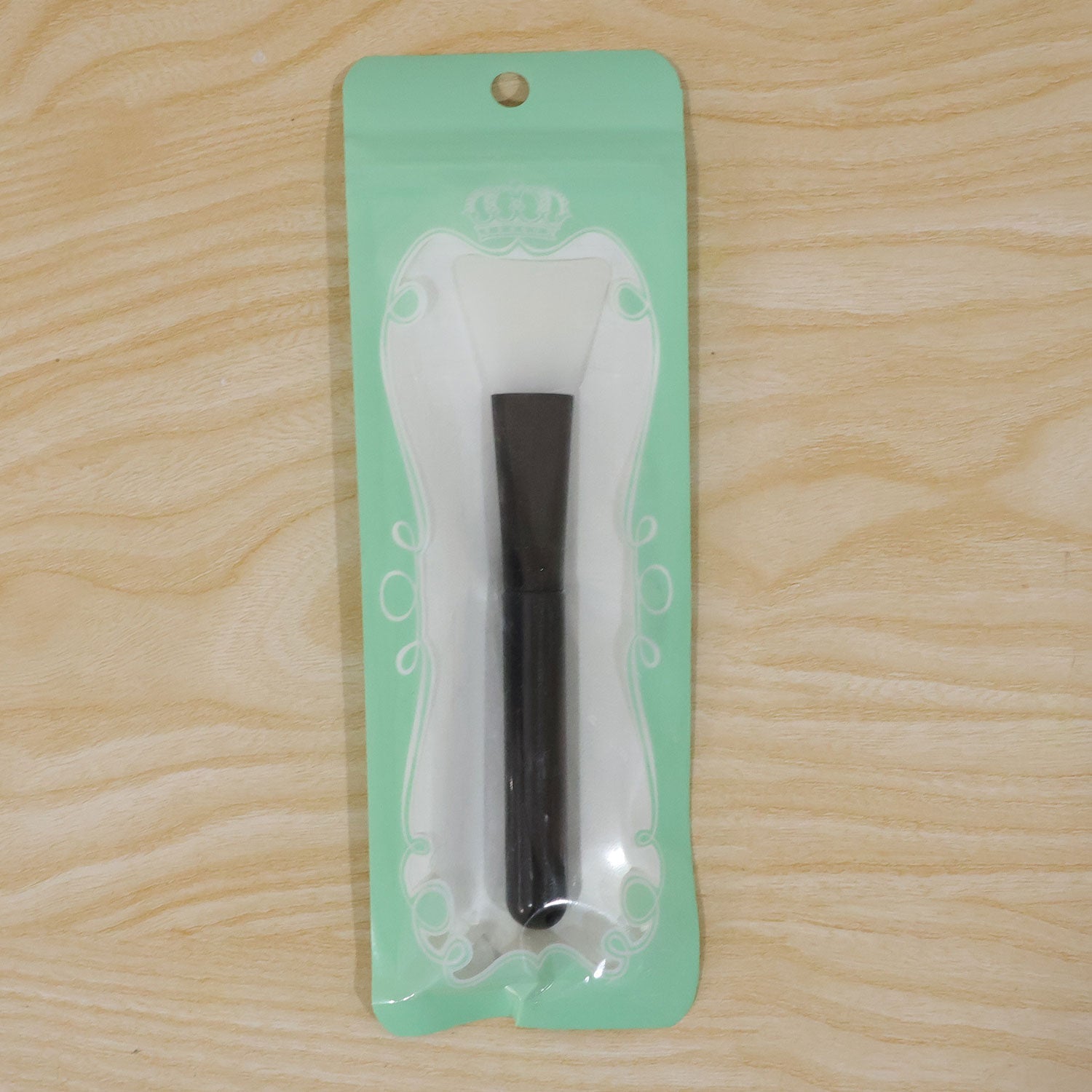 A black and white silicone mask brush is sealed inside its retail packaging, a clear plastic bag with a mint green backing,