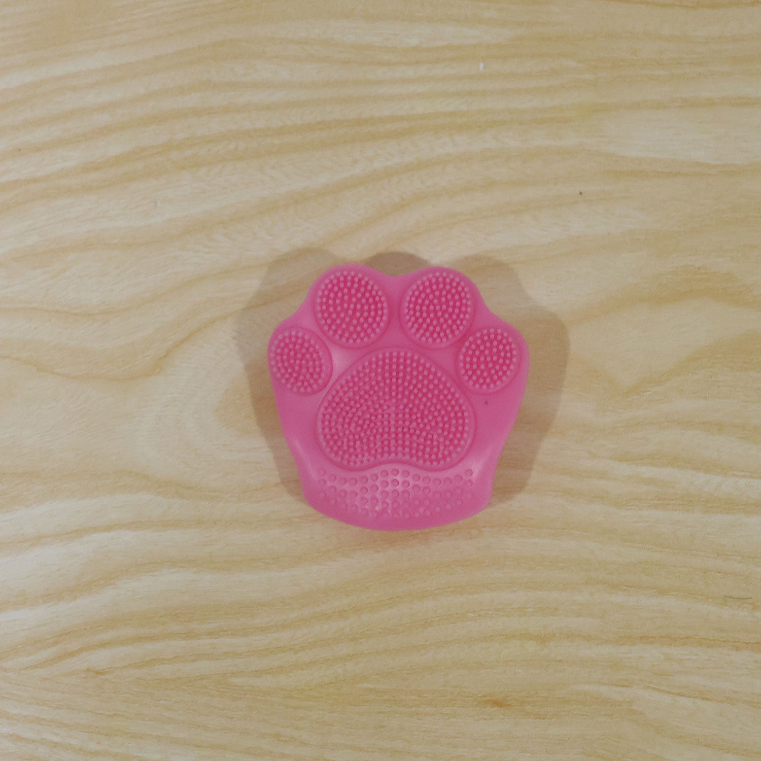 A single pink paw-shaped facial scrubber with silicone bristles rests on a light wooden table.