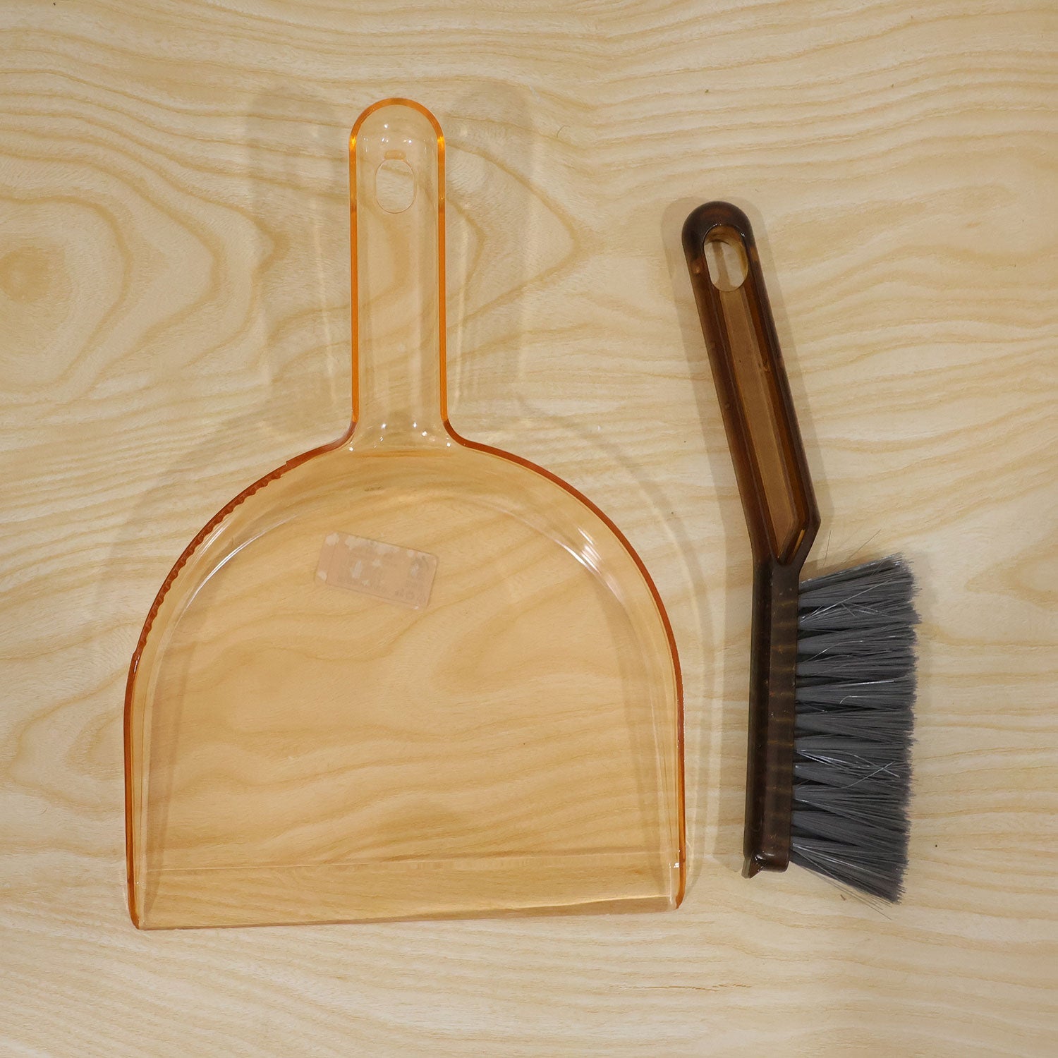 An amber plastic dustpan and a dark brown hand brush with gray bristles are placed on a light wooden surface.