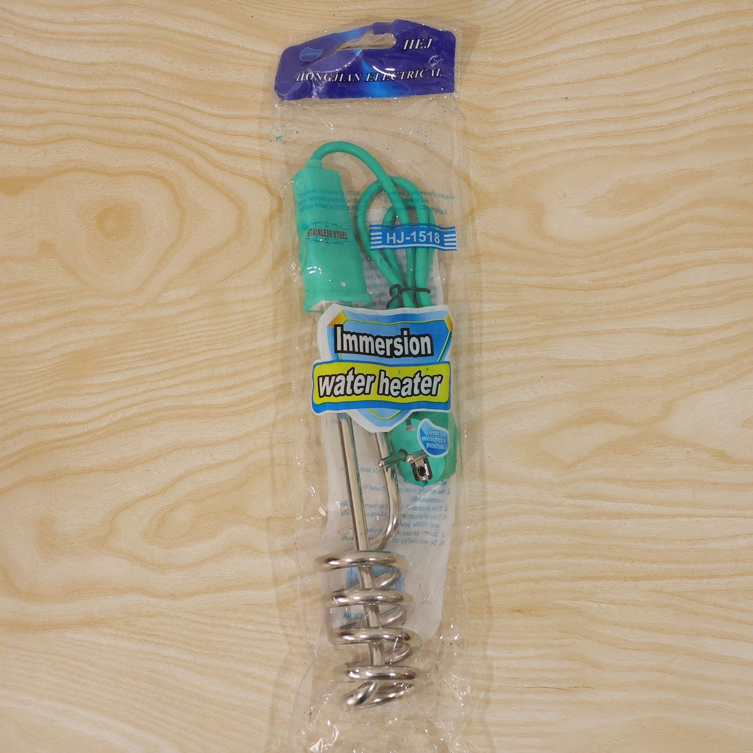 The teal and silver immersion water heater is sealed in clear plastic packaging with a label on a wooden surface.