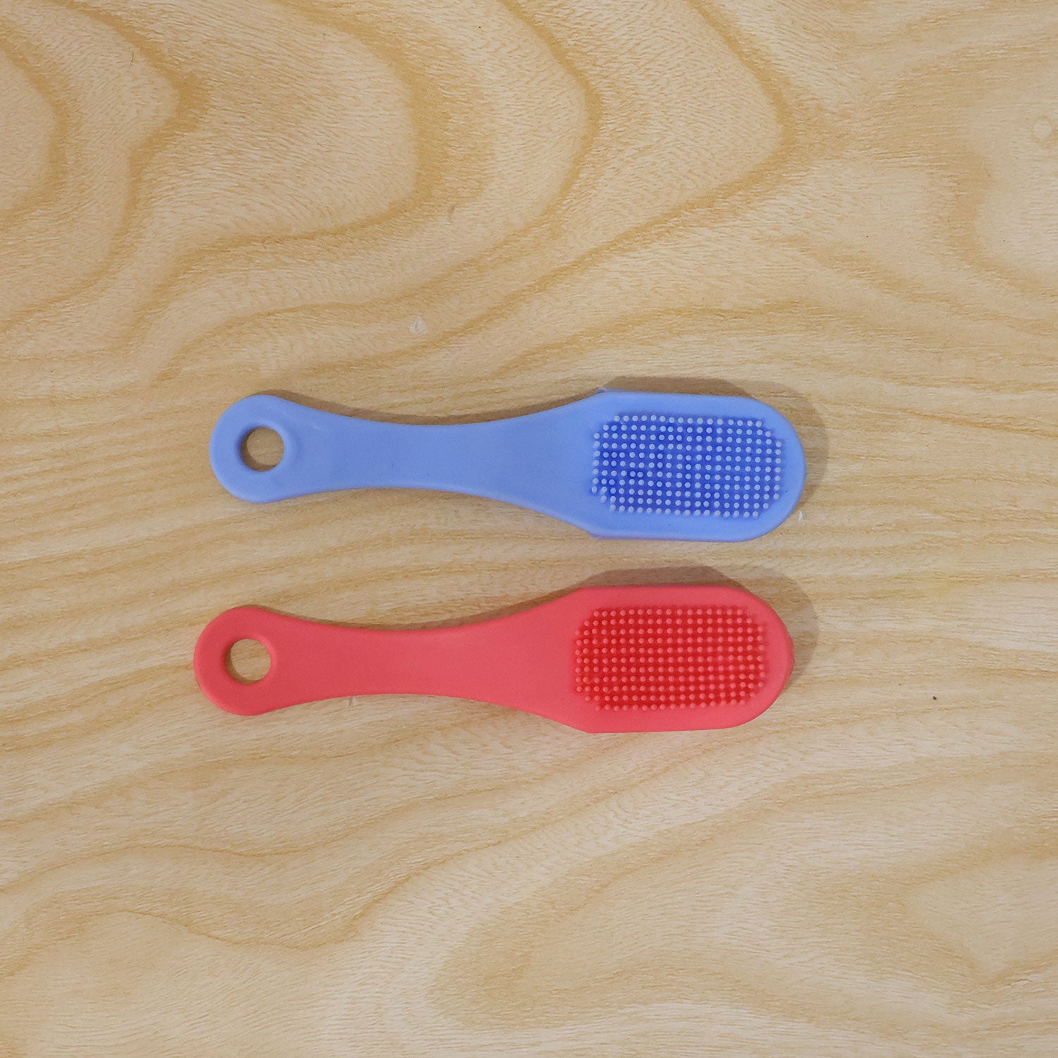 A blue and a pink silicone facial brush, both with textured bristles, lie flat on a light wooden surface.