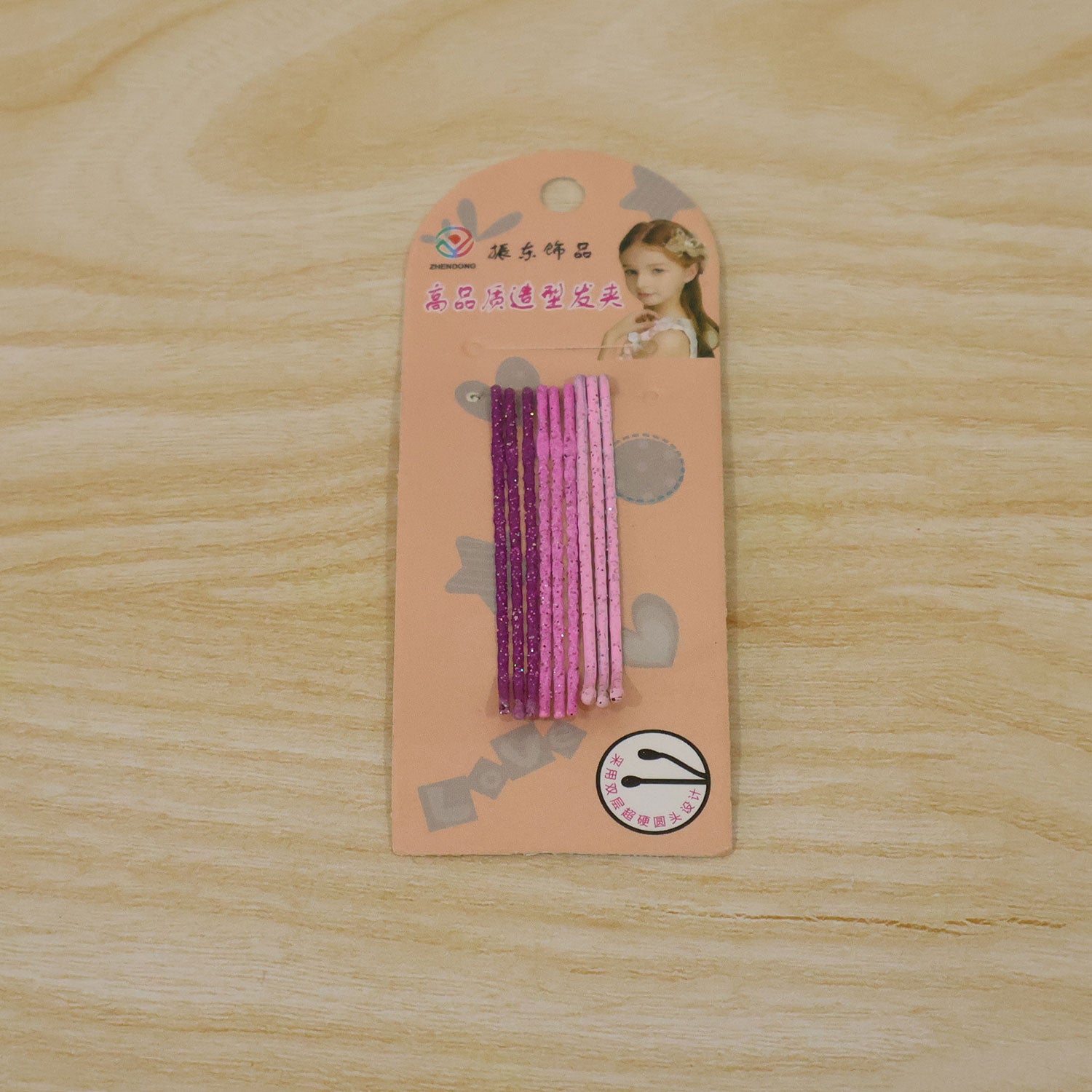 A retail package containing nine glitter bobby pins, five purple and four pink, rests on a light wooden surface.