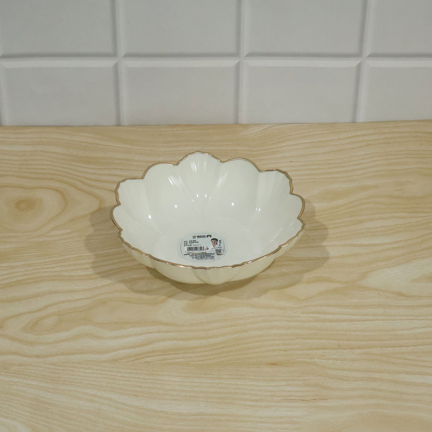 Empty white ceramic scalloped bowl with a gold rim on a wooden surface with a white tiled background.