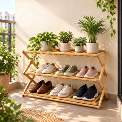 A light brown bamboo three-tier shoe rack holds various pairs of shoes and potted plants on a sunny balcony.