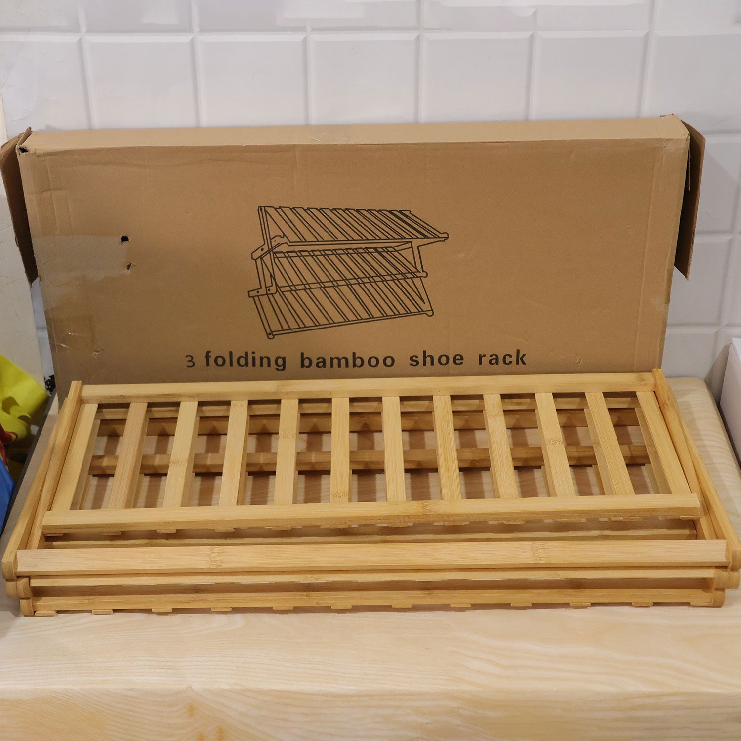 A folded bamboo shoe rack sits in front of its open cardboard packaging box, displaying the product's compact form.