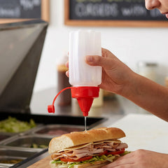 A person squeezes a white plastic bottle with a red cap, dispensing sauce onto a deli sandwich.