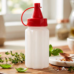 A small, empty, white ribbed plastic squeeze bottle with a bright red cap sits on a wooden board with herbs.
