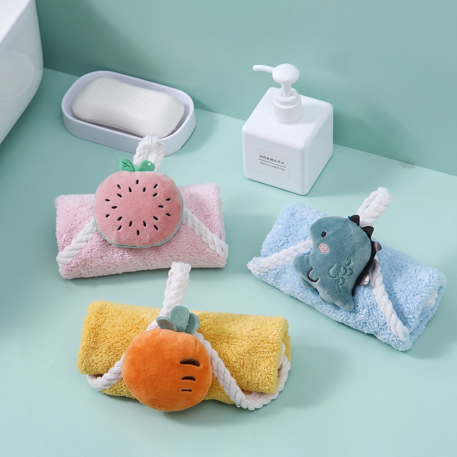 Three rolled colorful towels with plush fruit and dinosaur shapes, displayed on a teal surface next to bathroom accessories.
