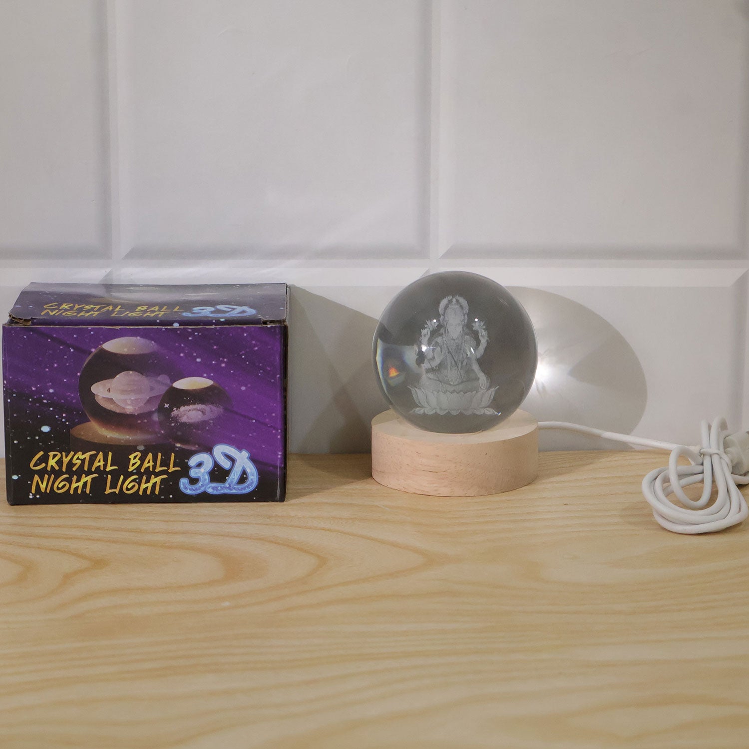 A crystal ball lamp with a Laxmi figure on a wooden base next to its purple and black packaging box.